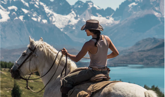 Woman wearing a leather hat rides a white horse with Patagonian mountains in the background.