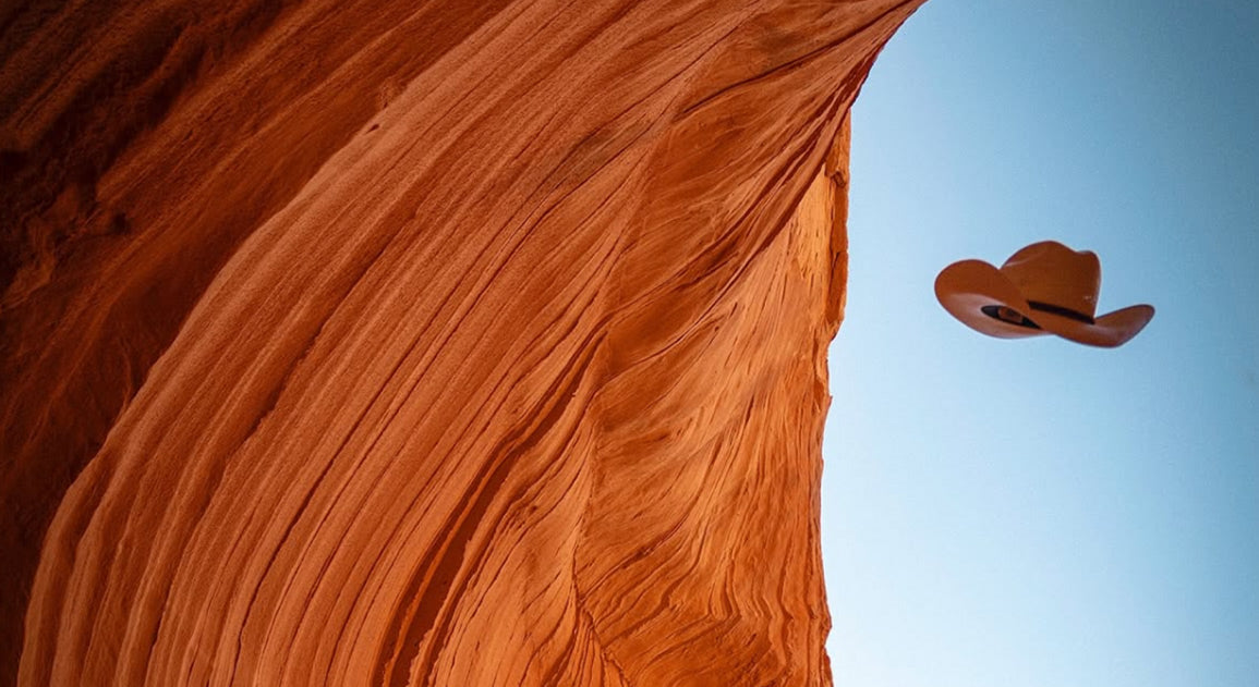 Tan cowboy hat tossed into a blue sky beside a curved red sandstone canyon wall