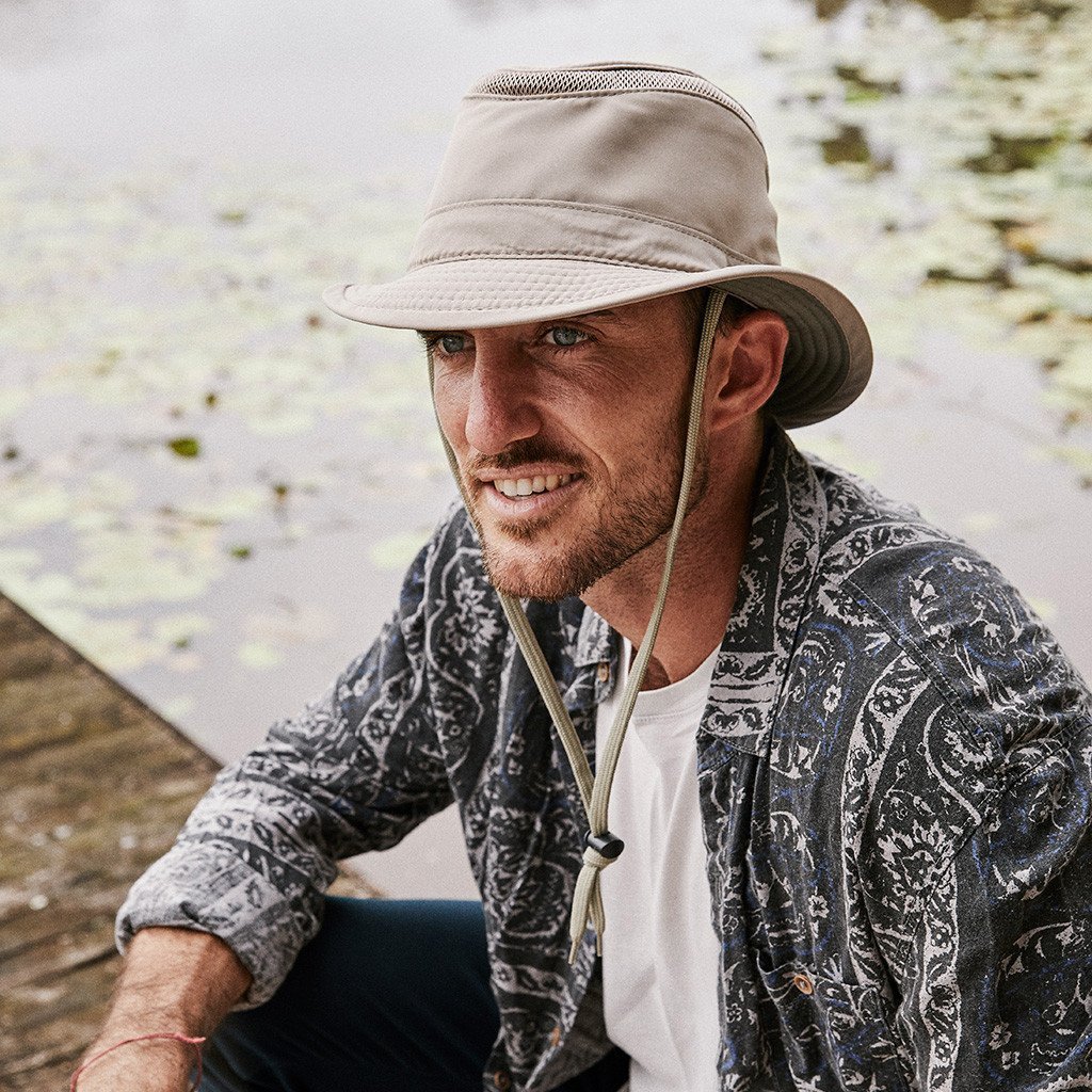 Man sitting on dock in recycled cloth hat in boating or sailing style with a thin air vent strip at top of crown and short fedora brim and chin cord in color Sand