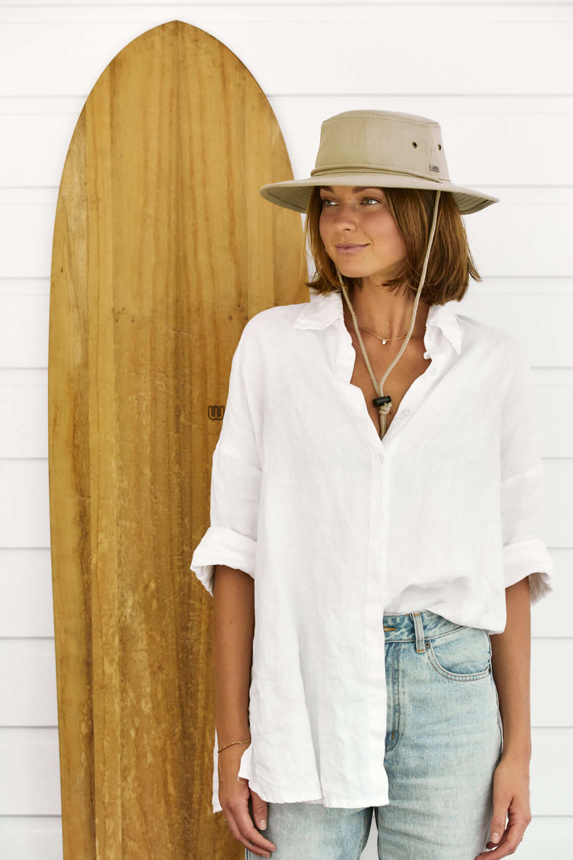 Woman standing next to a wooden surfboard in a Boonie bucket style hat made from organic cotton with wide sun protective brim and cotton chin cord