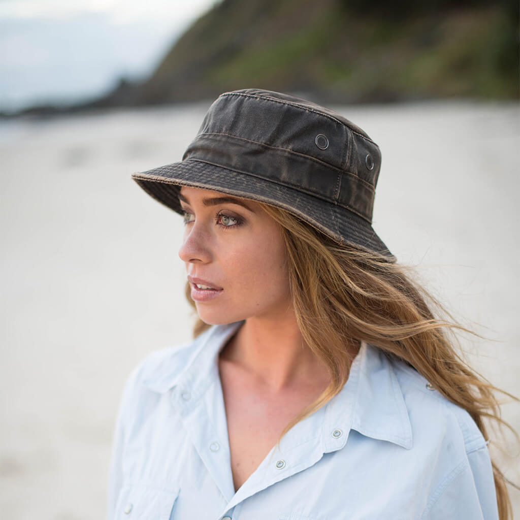 Woman on a beach in Byron Bay wearing waterproof cloth bucket Hat with brass colored mesh eyelets and Conner Hat emblem