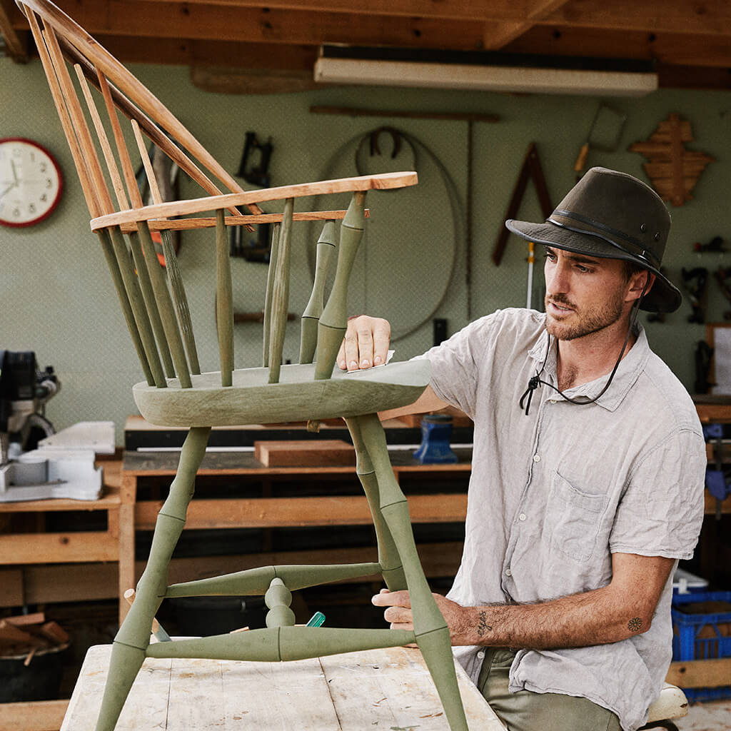 Man in wood shop wearing Australian outback oilskin cotton hat with leather chin cord and brass colored mesh eyelets