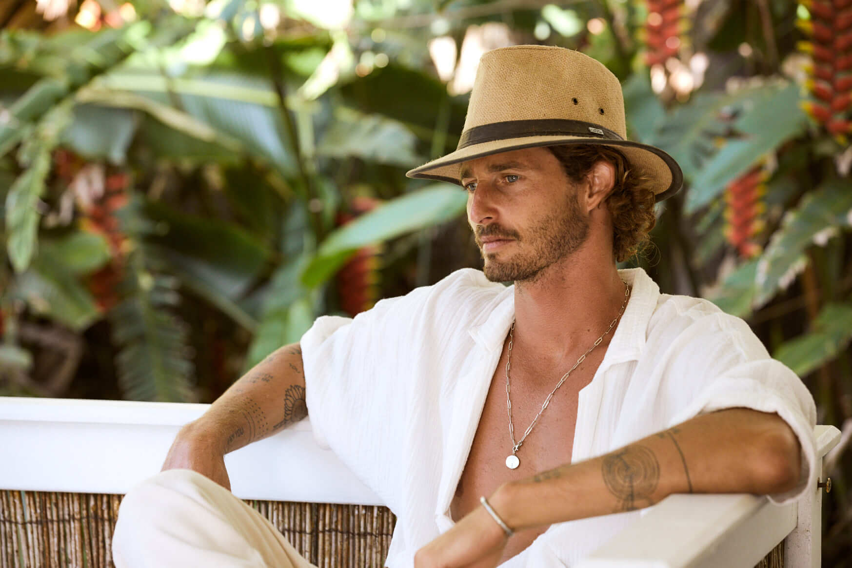 Man sitting in tropical location wearing safari style pinched front sun protection hat made from natural colored hemp with a dark brown band and trim