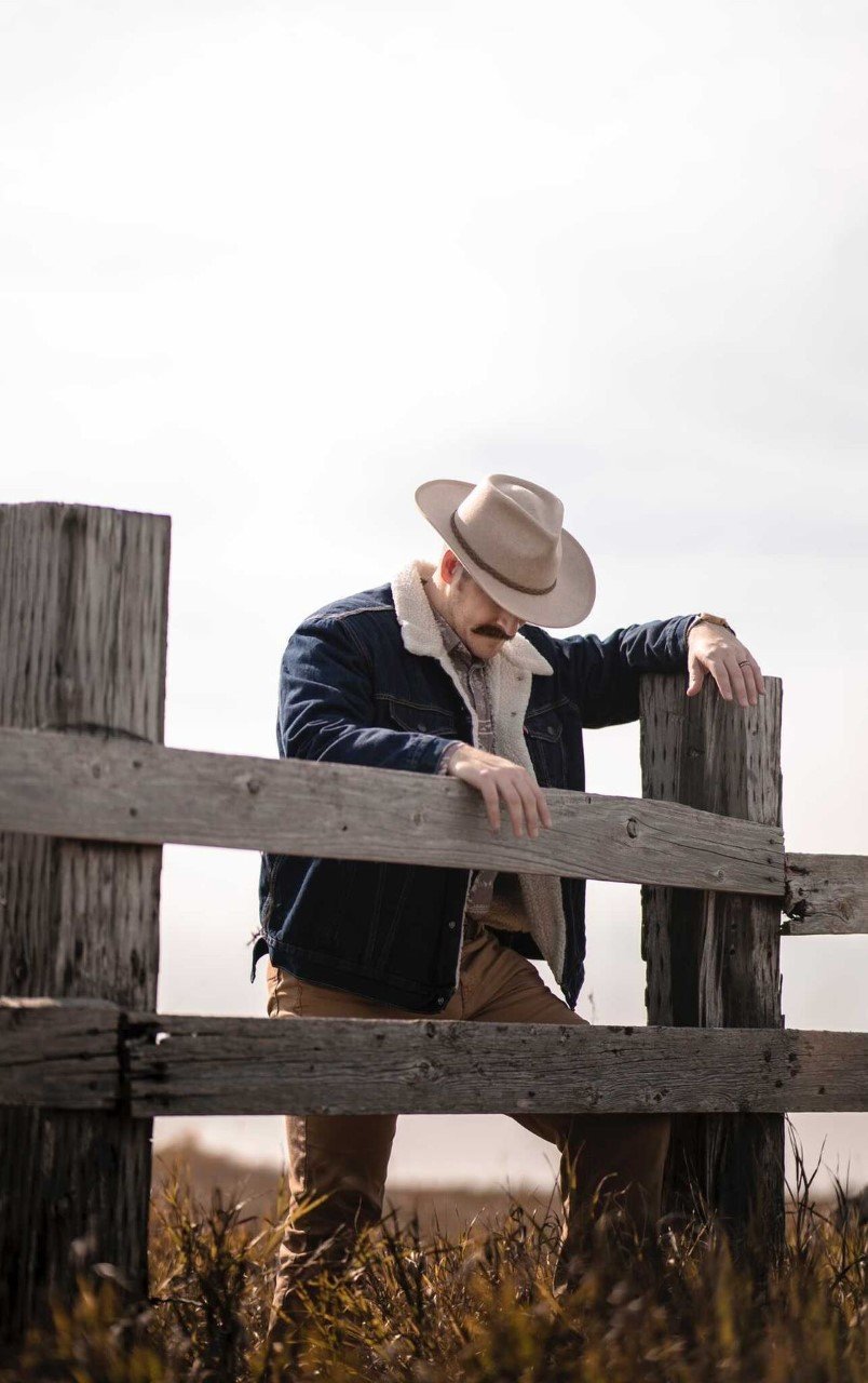 Man standing on wooden fence line in Americana style clothes wearing outback style hat with stiff brim and bound edge with a braided vegan band