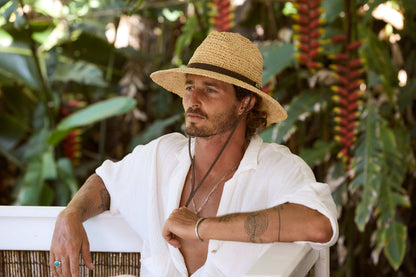 Man sitting in tropical setting wearing wide brim sun protection raffia straw hat hand crocheted in natural color with Brown hat band and Brown chin cord