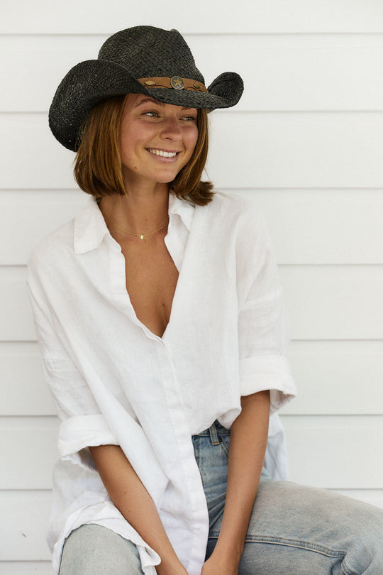 Woman sitting on chair smiling wearing Conner Hats Country Western straw hat hand braided from raffia in Black color with faux leather band and brass colored star and conchos