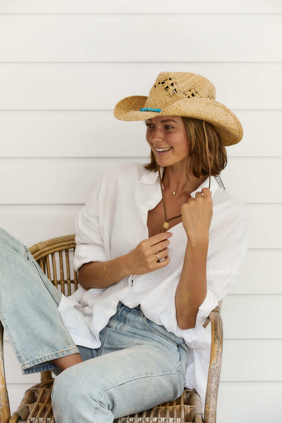 Woman lounging on chair wearing organic raffia western straw hat in color Natural with turquoise colored stones and vented crown for air flow and features a faux leather chin cord