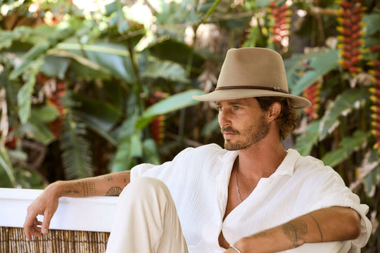 Man sitting outside in Putty wool outback hat with thin tooled leather hat band and Conner emblem with tropical flowers in the background 